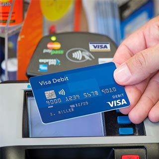 Person holding a Visa credit card in front of a payment terminal displaying the contactless symbol and credit card logos.
